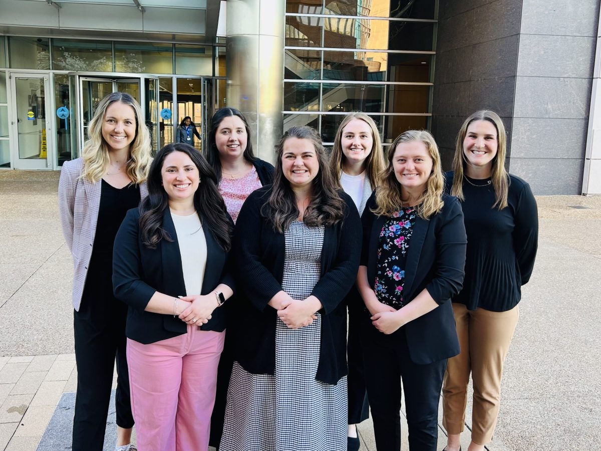 A group of seven smiling VUSM students and faculty poses for a photo outside a modern building.