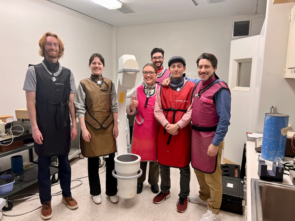 Medical physics students in a lab, wearing protective lead vests and smiling for the camera.