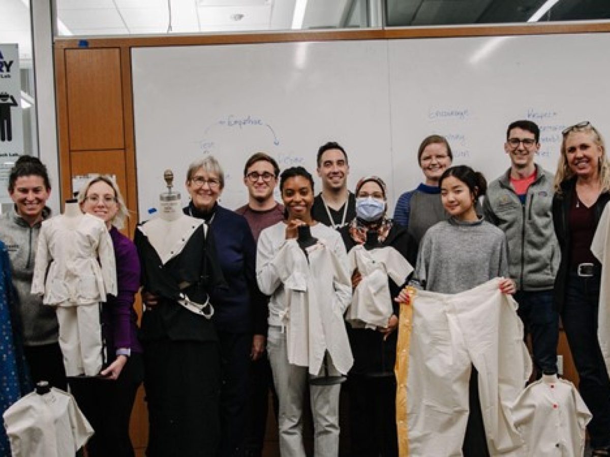 A group of students and faculty smile and pose with garment-making projects in a classroom.