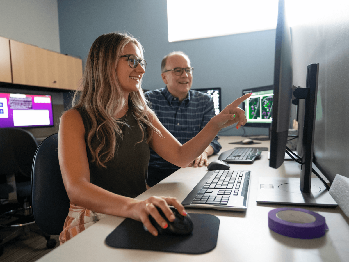 A student sitting at a desk in front of a computer and a faculty member sitting next to them. They look to be discussing what's on a computer screen.